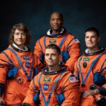 The four Artemis II astronauts—Christina Koch, Reid Wiseman, Victor Glover, and Jeremy Hansen—standing together in their dark blue flight suits against a black background.