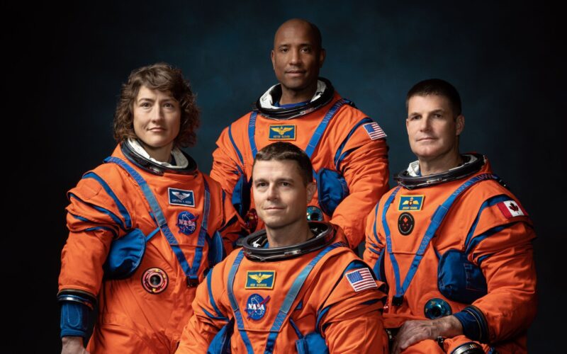 The four Artemis II astronauts—Christina Koch, Reid Wiseman, Victor Glover, and Jeremy Hansen—standing together in their dark blue flight suits against a black background.