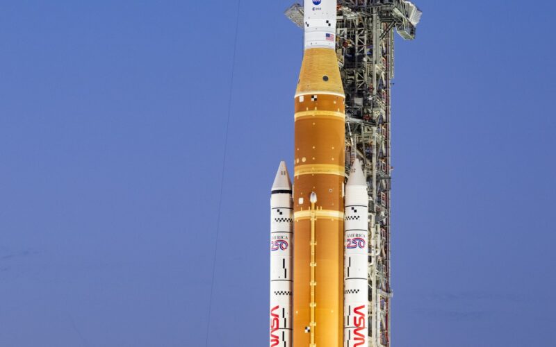 A towering orange and white NASA SLS rocket and Orion capsule stand vertically on a mobile launcher at night, brightly illuminated by spotlights against a dark sky at Launch Pad 39B.
